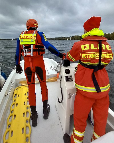 Auf diesem Bild sieht man die wasserseitige Absicherung einer Regatta. Vorne auf dem Boot befindet sich ein Rettungsschwimmer in Schutzausrüstung, der bereit ist, ins Wasser zu gehen. Am Steuerstand des Boots steht der Bootsführer.